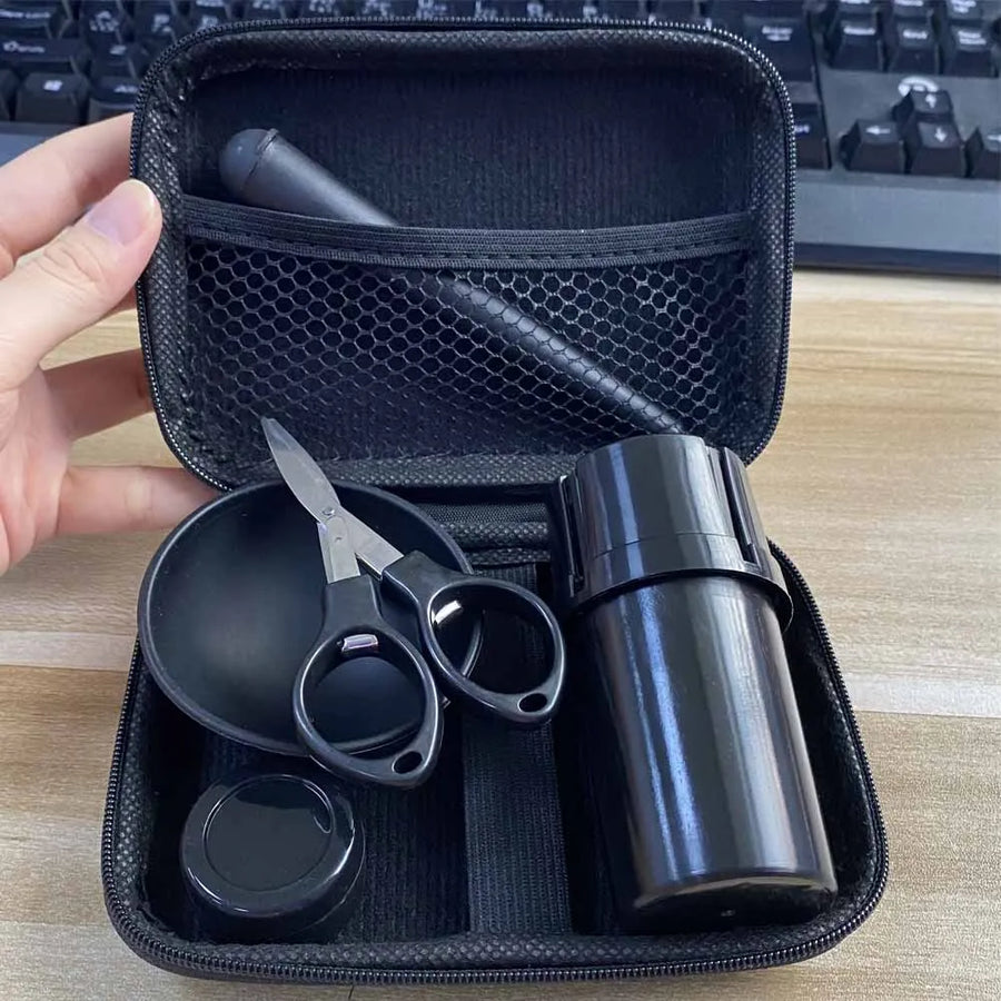 A black zippered carrying case opened to show a travel coffee kit inside, including a black travel mug, scissors, a small container, and a pen, held by a hand on a desk with a keyboard in the background.